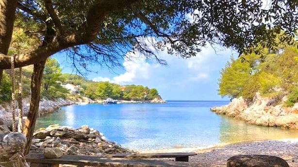 Un paisaje costero tranquilo con un banco de madera y un montículo de piedras, con vista a un mar en calma y un litoral rocoso.
