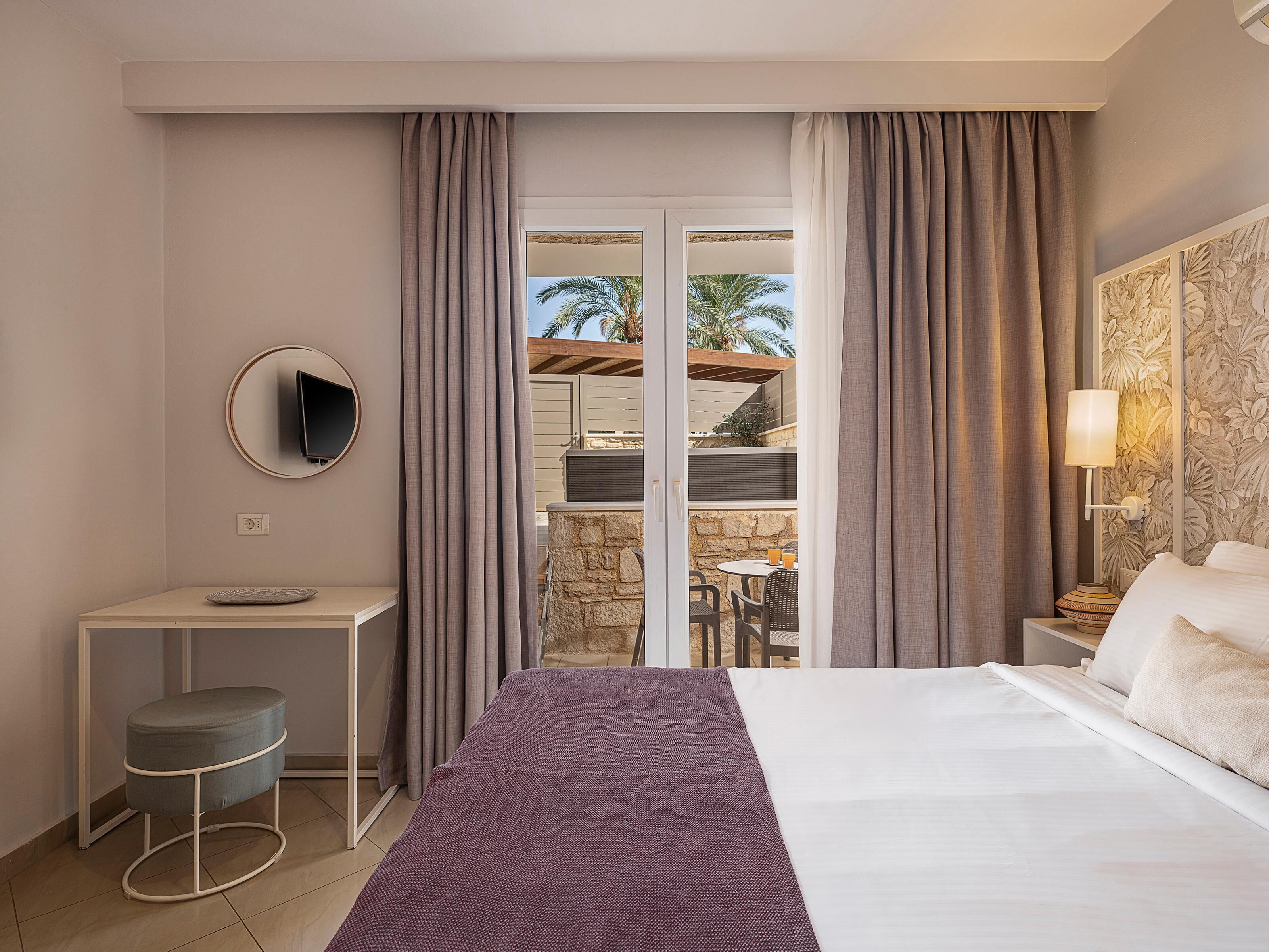 Habitación de hotel con cama, escritorio con silla, un taburete pequeño, televisor montado en la pared y vista al área exterior a través de una puerta de vidrio.