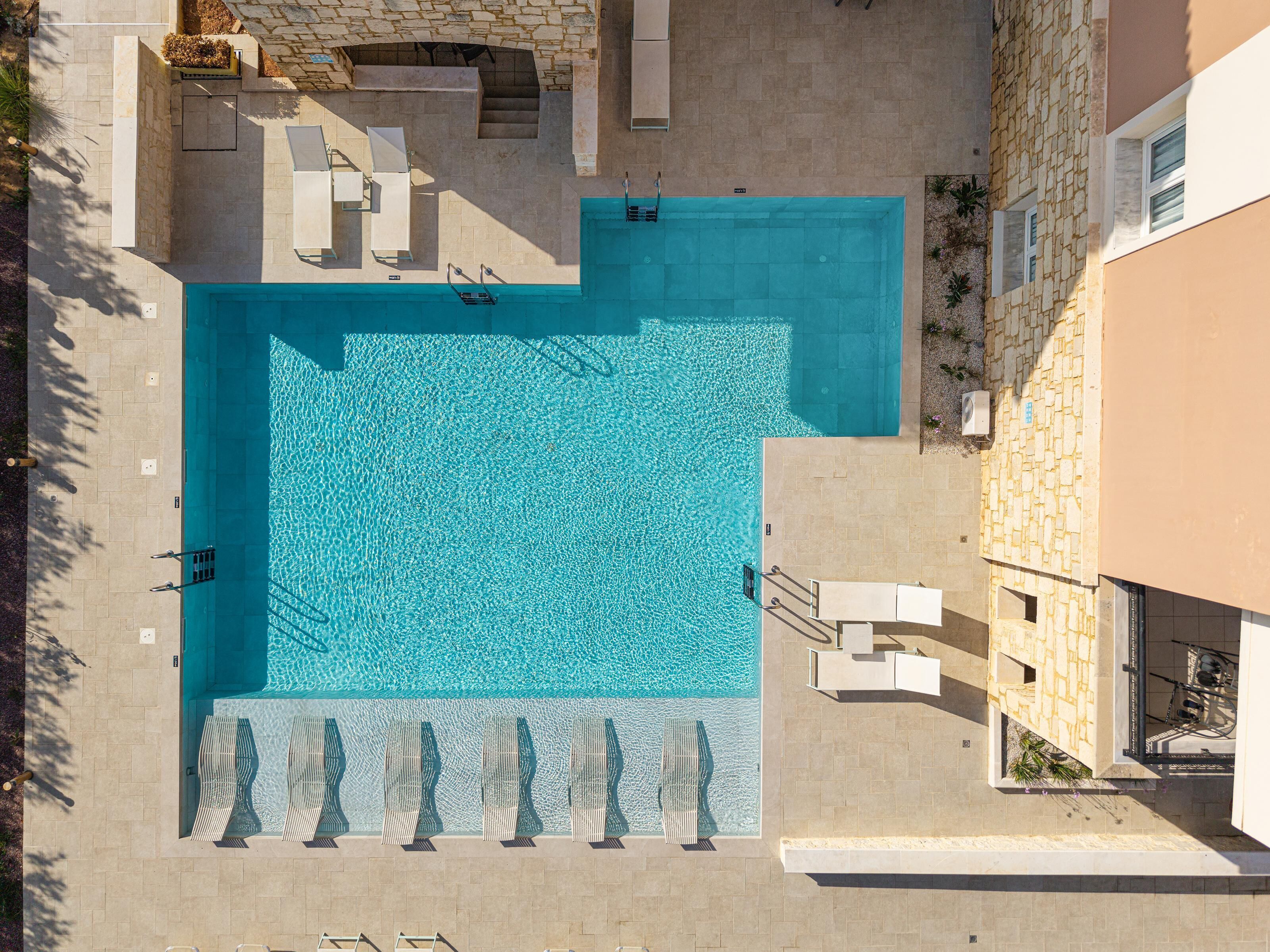 Eine Vogelperspektive auf einen Pool mit Fliesen, Liegestühlen und einer Steinmauer.