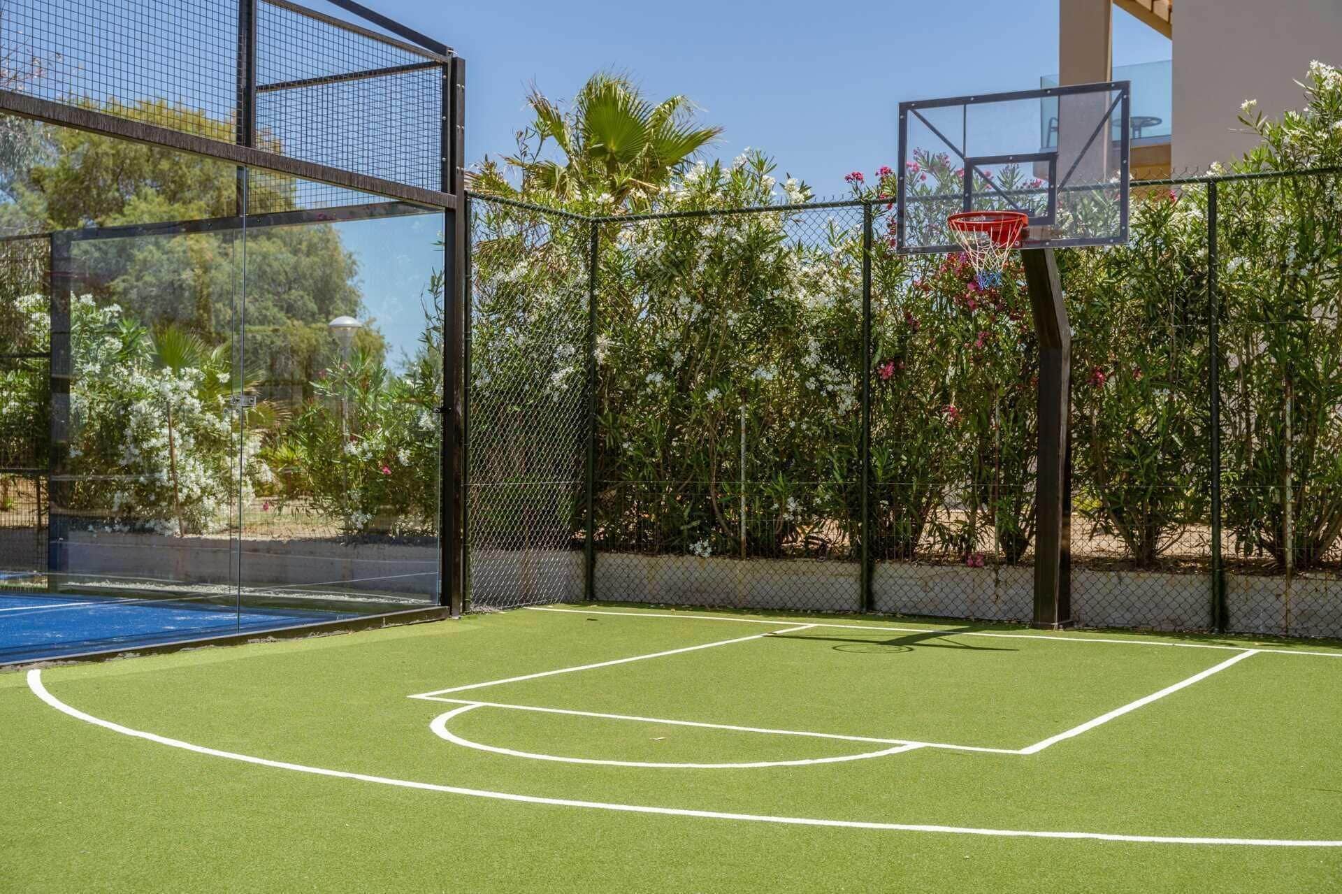 An outdoor basketball court with a chain-link fence, surrounded by flowering plants and trees.