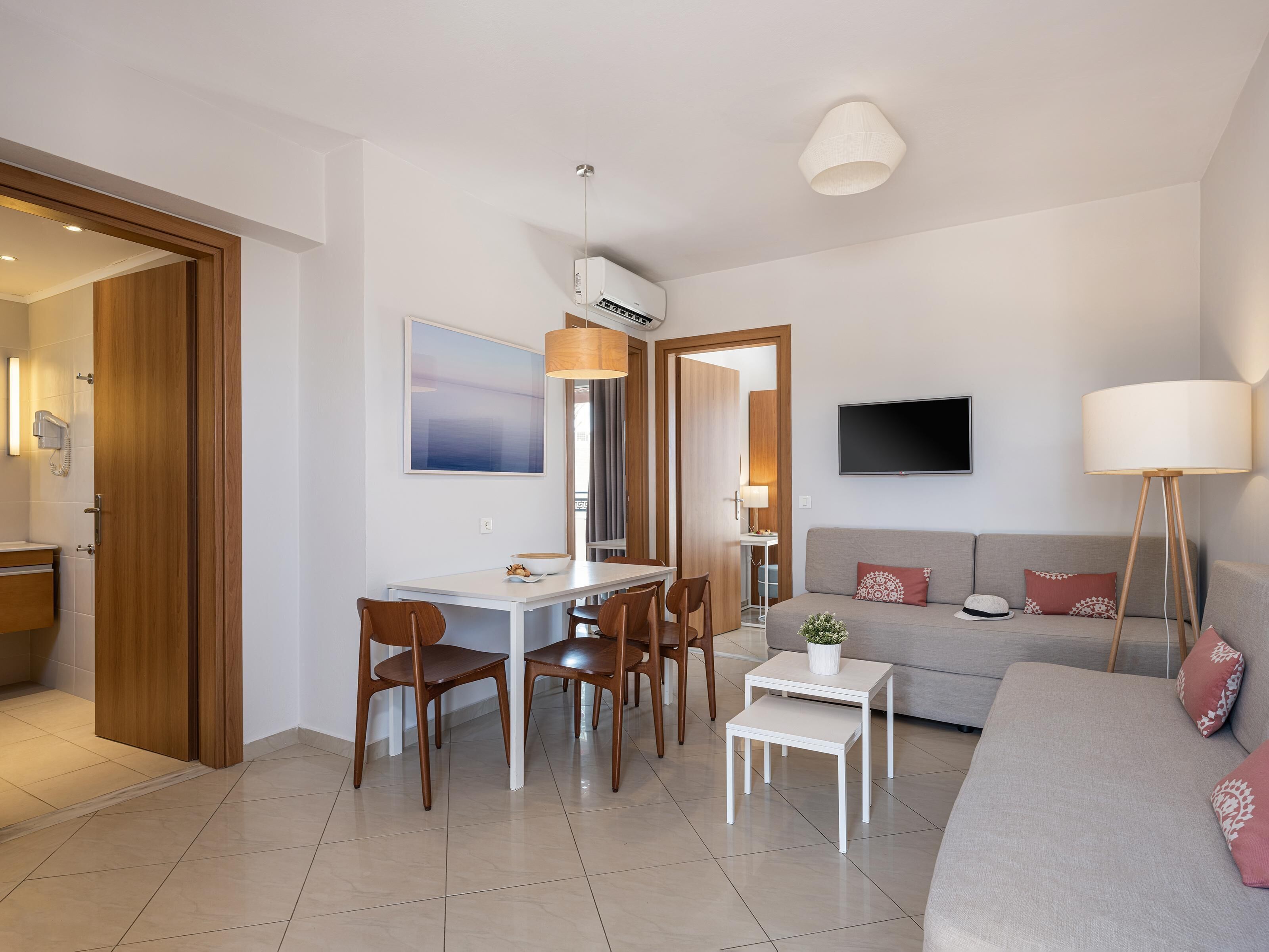 A modern living room with a sofa, a dining table with chairs, a TV mounted on the wall, and a lamp.