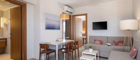 A modern living room with a sofa, a dining table with chairs, a TV mounted on the wall, and a lamp.