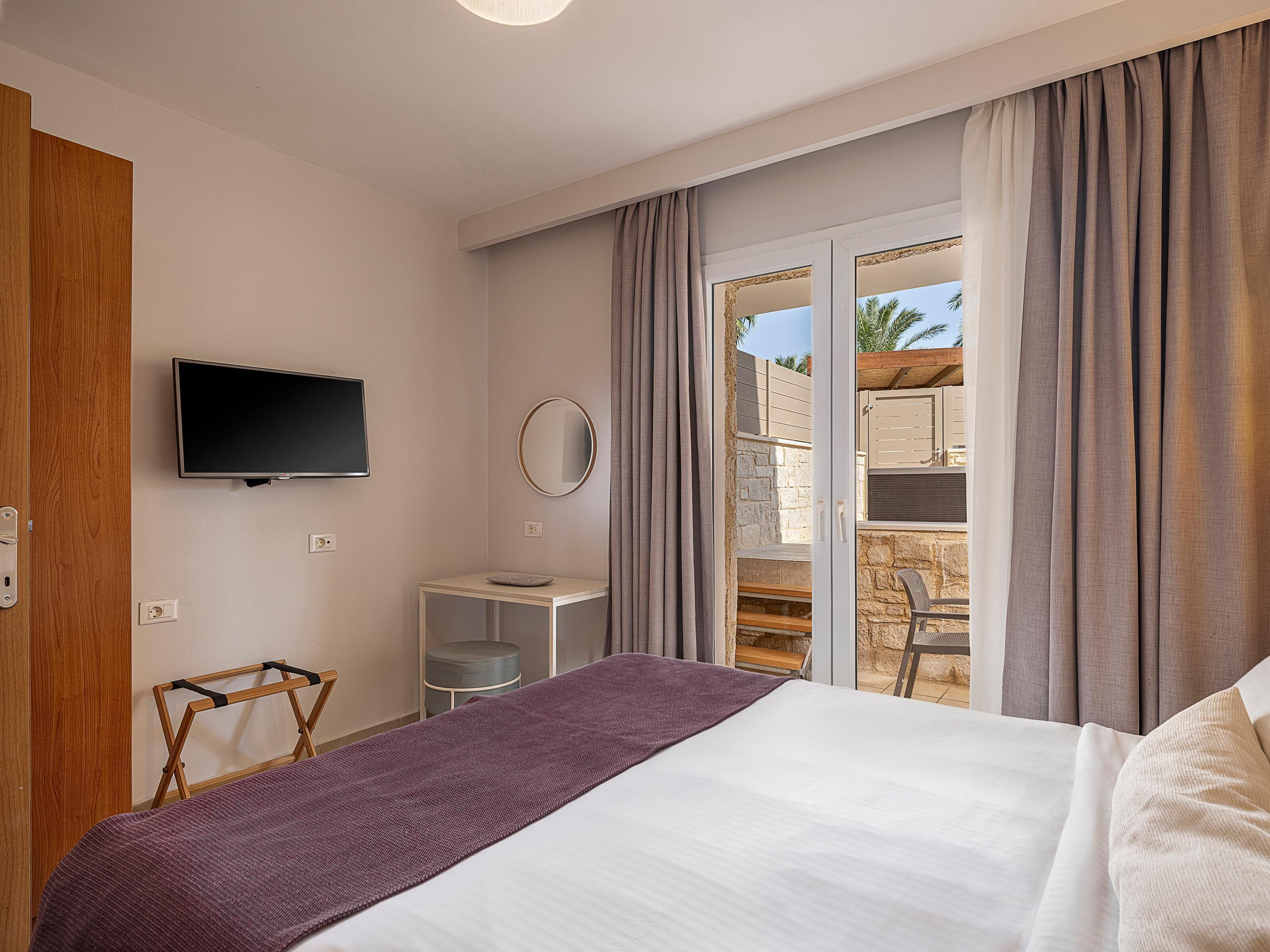 A hotel room with a bed, a TV, a small table, a mirror, and a view of the outdoor area.