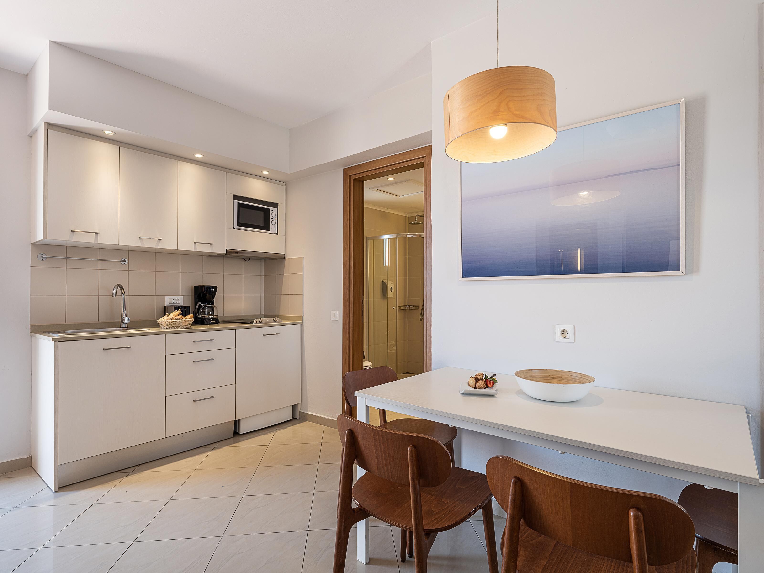 A modern kitchen with white cabinets, a microwave, and a coffee maker. There is a dining table with chairs and a large painting on the wall.