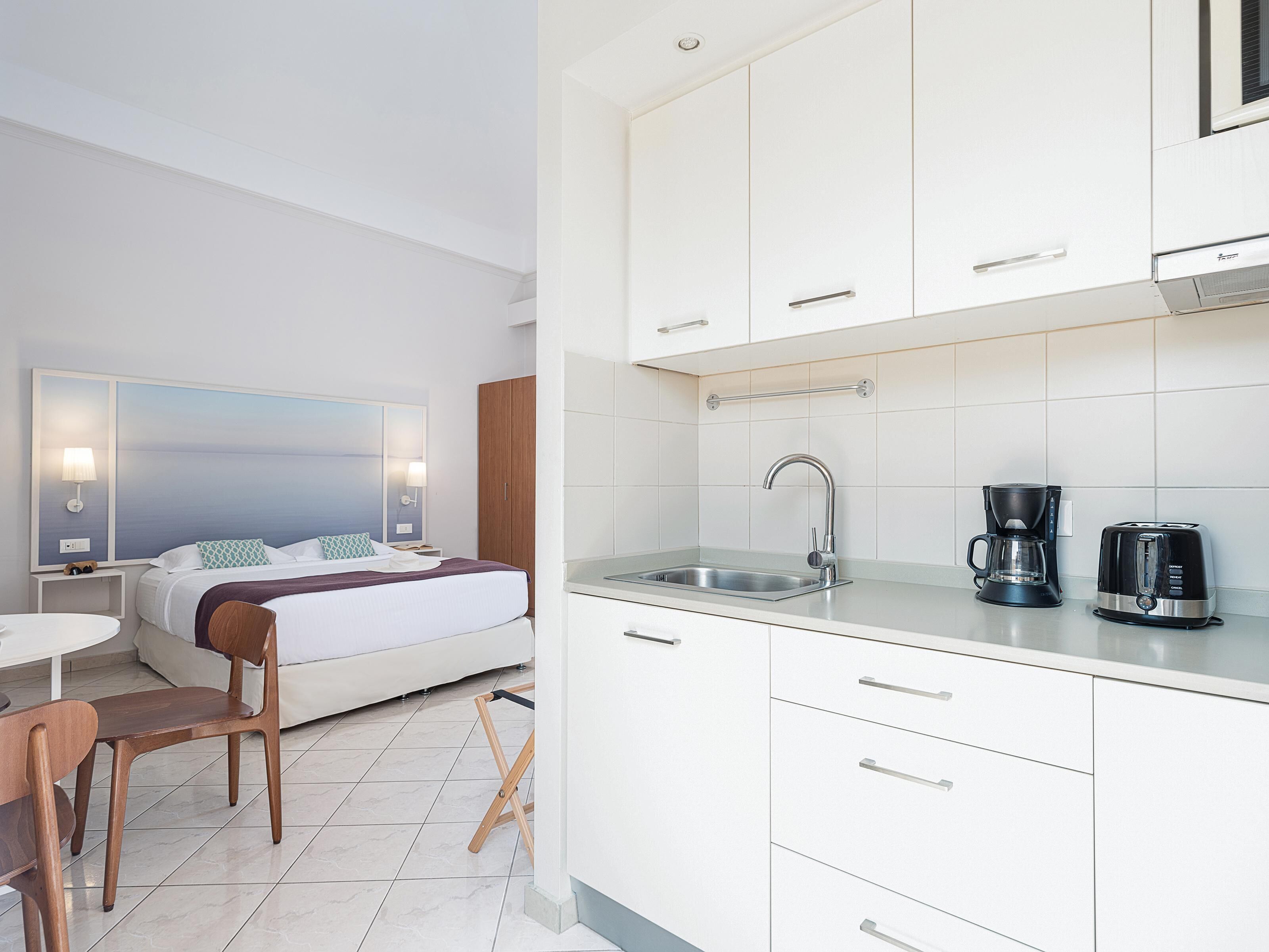 Una habitación de hotel moderna con cama, mesita de noche, lámpara y vistas al mar. La habitación cuenta con una kitchenette equipada con fregadero, cafetera y tostadora.