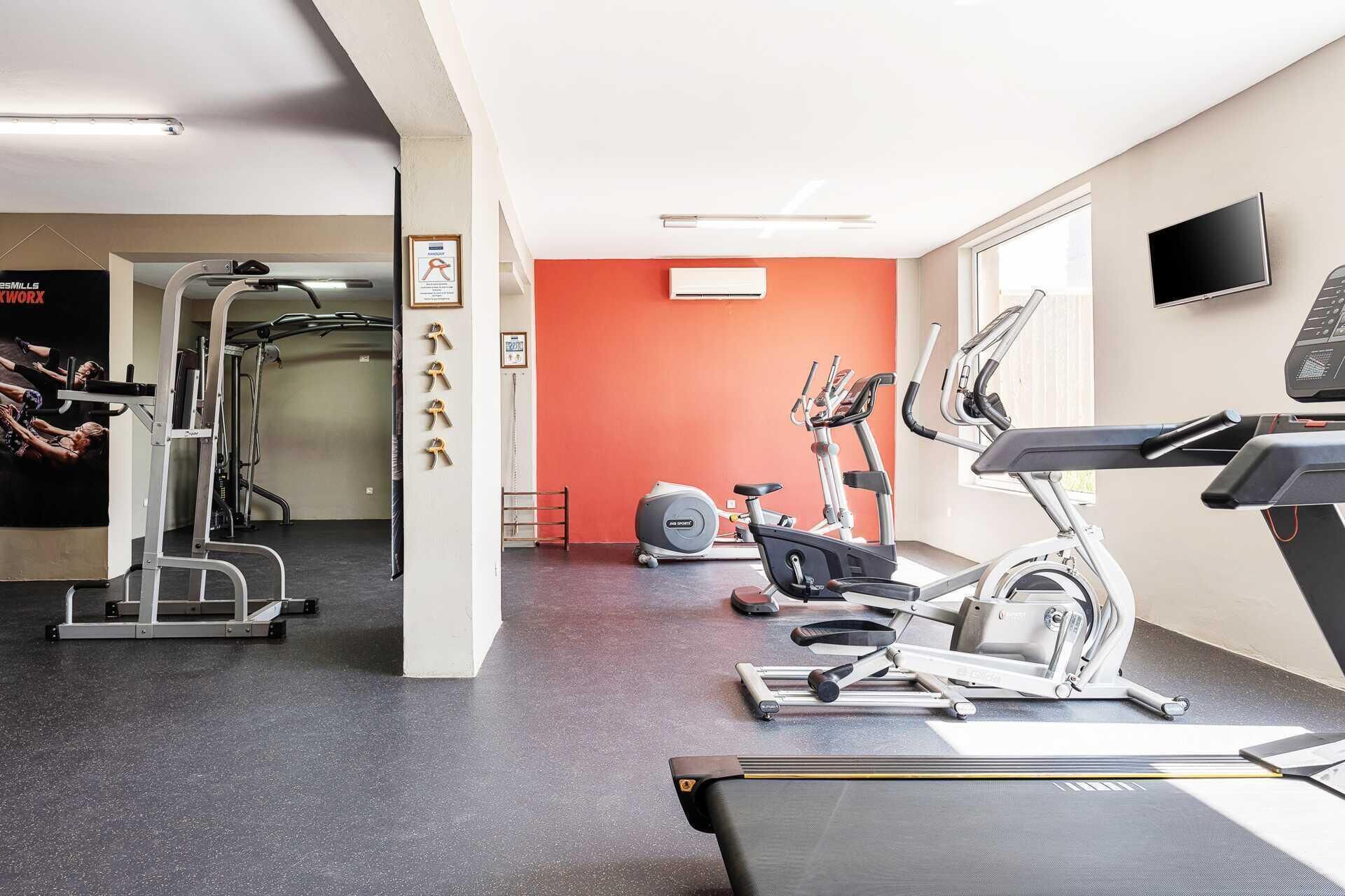 Et fitnesscenter med forskellig træningsudstyr, heriblandt løbebånd og styrkemaskiner, samt en rød accentvæg.