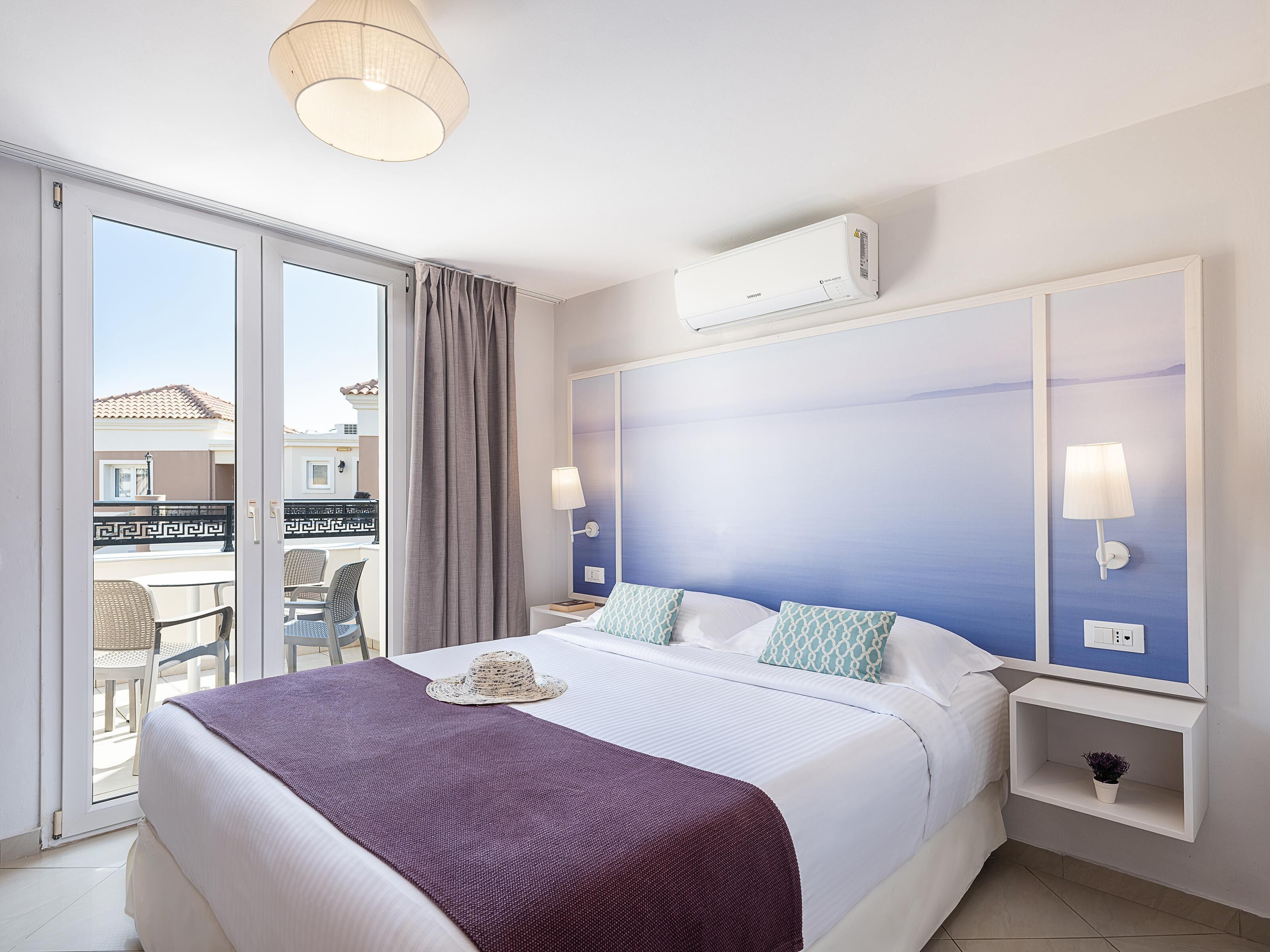 Habitación de hotel con una cama grande, ventilador de techo, ventana con cortinas, mesita de noche con lámpara y vistas al balcón exterior.