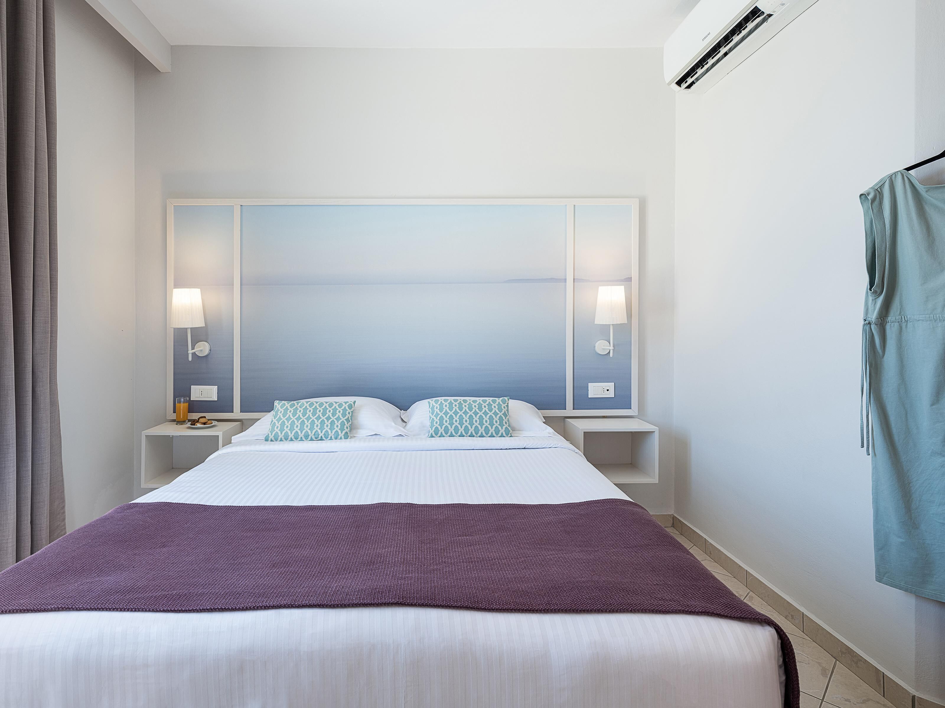 Een opgemaakt bed met een paarse deken, een grote tv aan de muur en twee bedlampen.