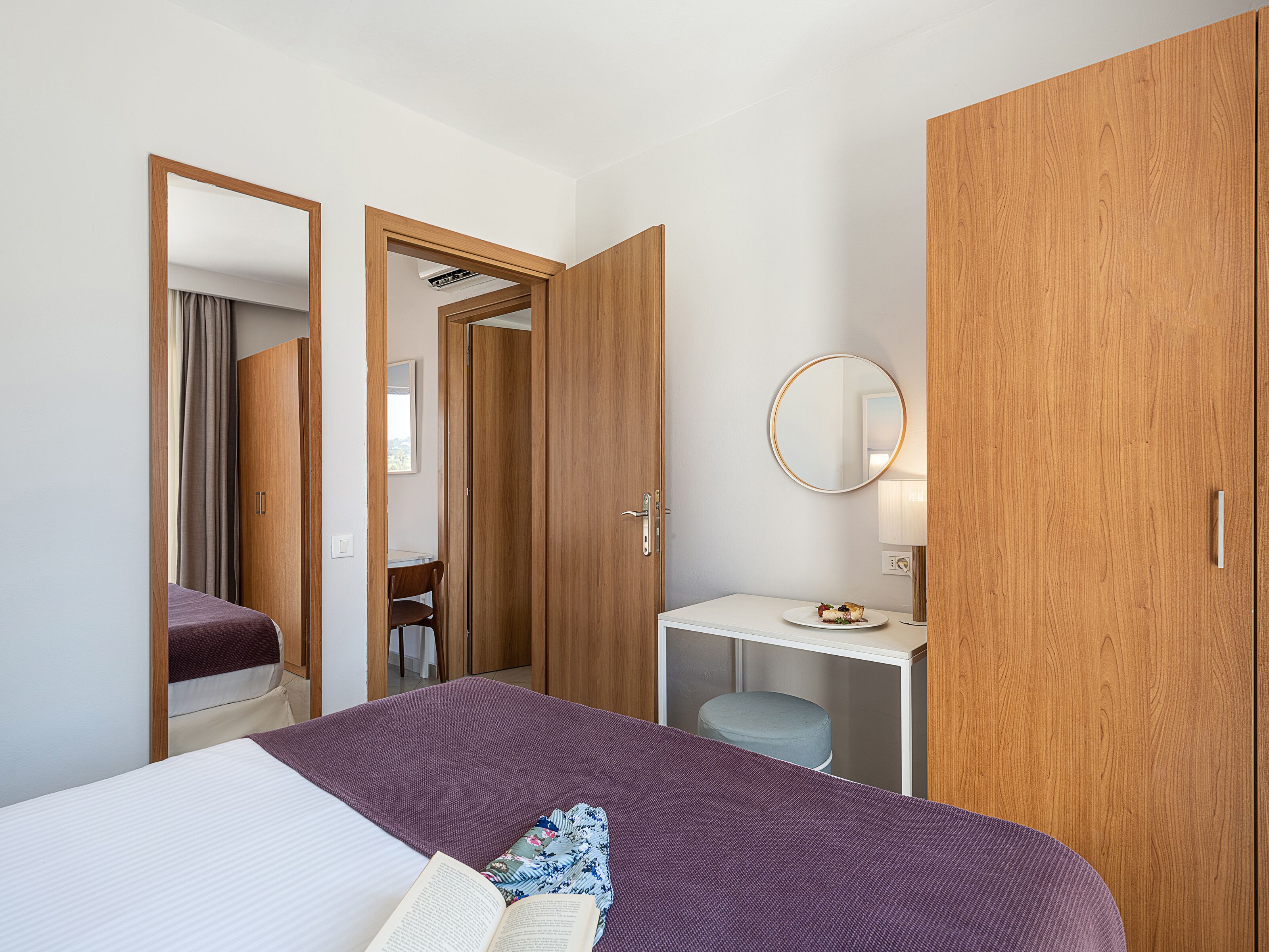 Habitación de hotel con cama, escritorio, silla, espejo redondo y armarios de madera.