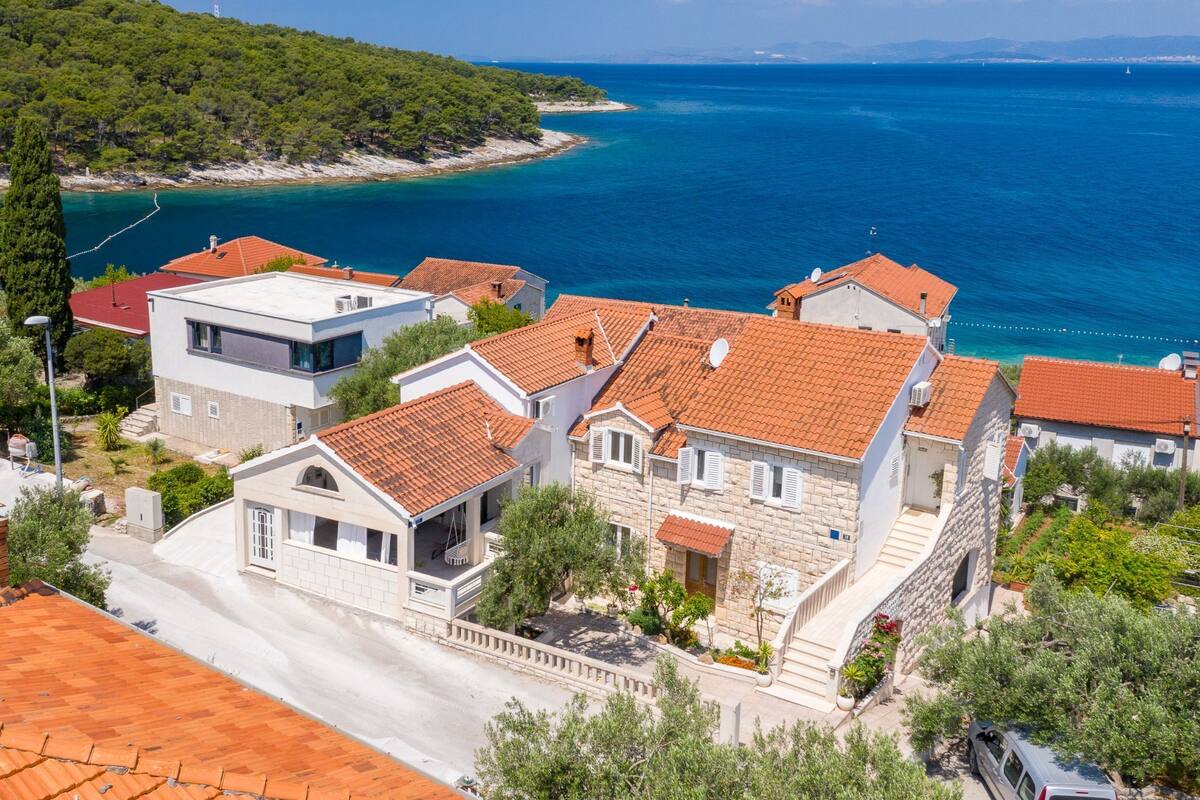 A coastal residential area with houses featuring red-tiled roofs and stone walls, overlooking a clear blue sea.