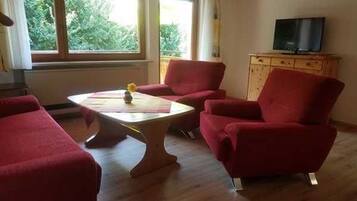 Ein Zimmer mit Holzboden, einem runden Tisch mit einer Vase, zwei roten Sesseln, einem Fernseher auf einer Holzkommode und einem Fenster mit Blick auf Grünflächen.