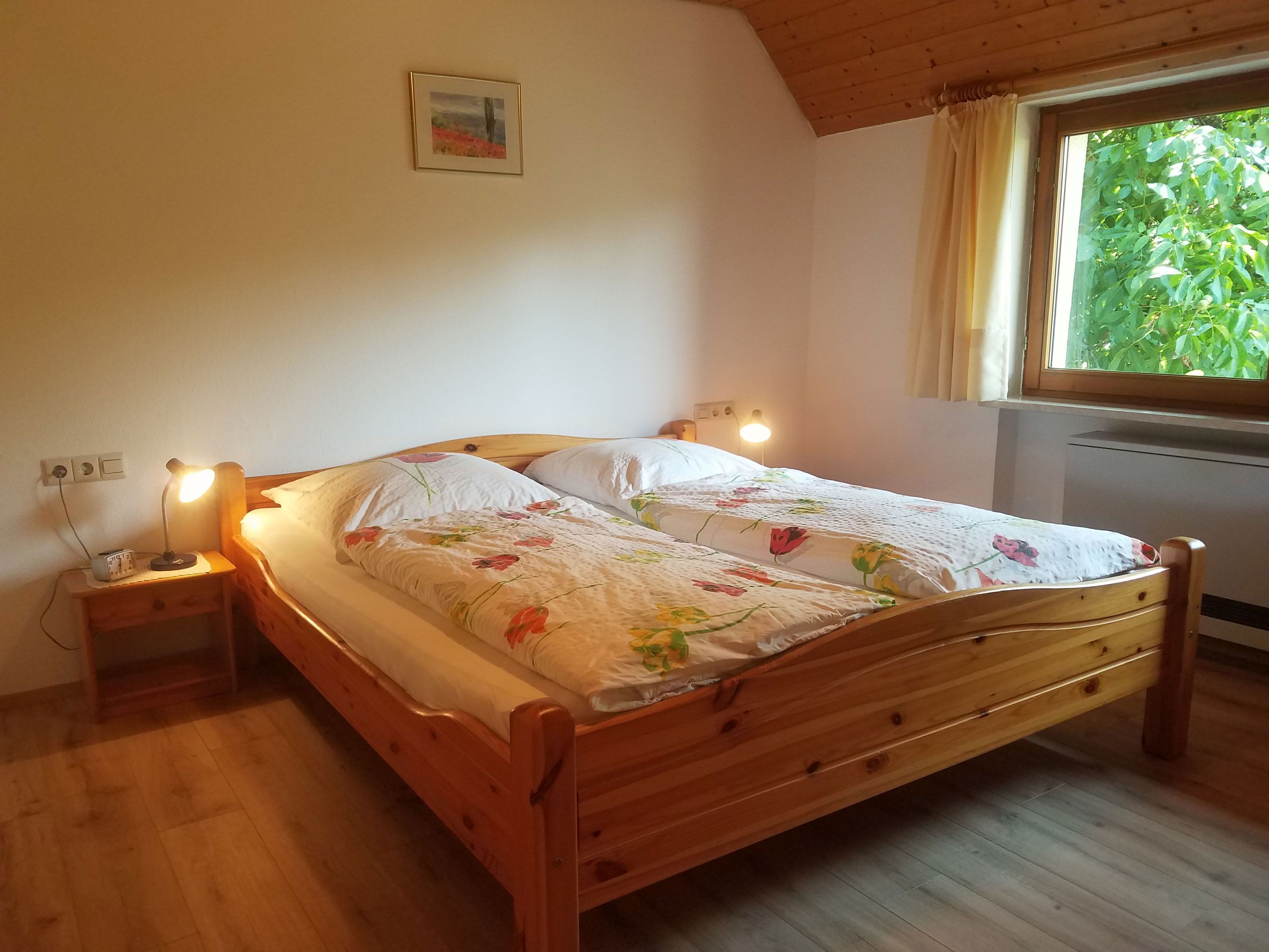Ein hölzernes Bett mit floralem Überwurf, zwei Nachttischlampen, ein Nachttisch, eine Wandsteckdose und ein Fenster mit Vorhängen.