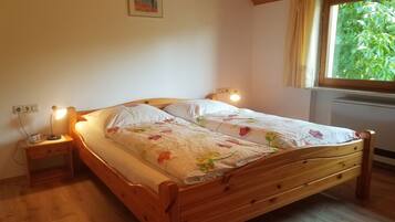 Ein hölzernes Bett mit floralem Überwurf, zwei Nachttischlampen, ein Nachttisch, eine Wandsteckdose und ein Fenster mit Vorhängen.