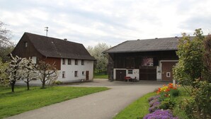 A rural setting with a white house and a barn, a paved driveway, and flowering plants.