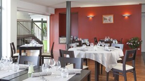 A restaurant with multiple tables set for dining, a staircase leading to an upper level, and a red wall with two wall-mounted lights. - Gite in the heart of the village (Belleville-sur-Loire)