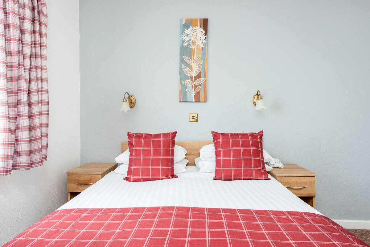 A neatly made bed with a red and white checkered bedspread, two red pillows, and a white pillow. There is a wooden nightstand with a white phone and a small decorative item on it. A wall-mounted light fixture is visible on the left side of the bed.
