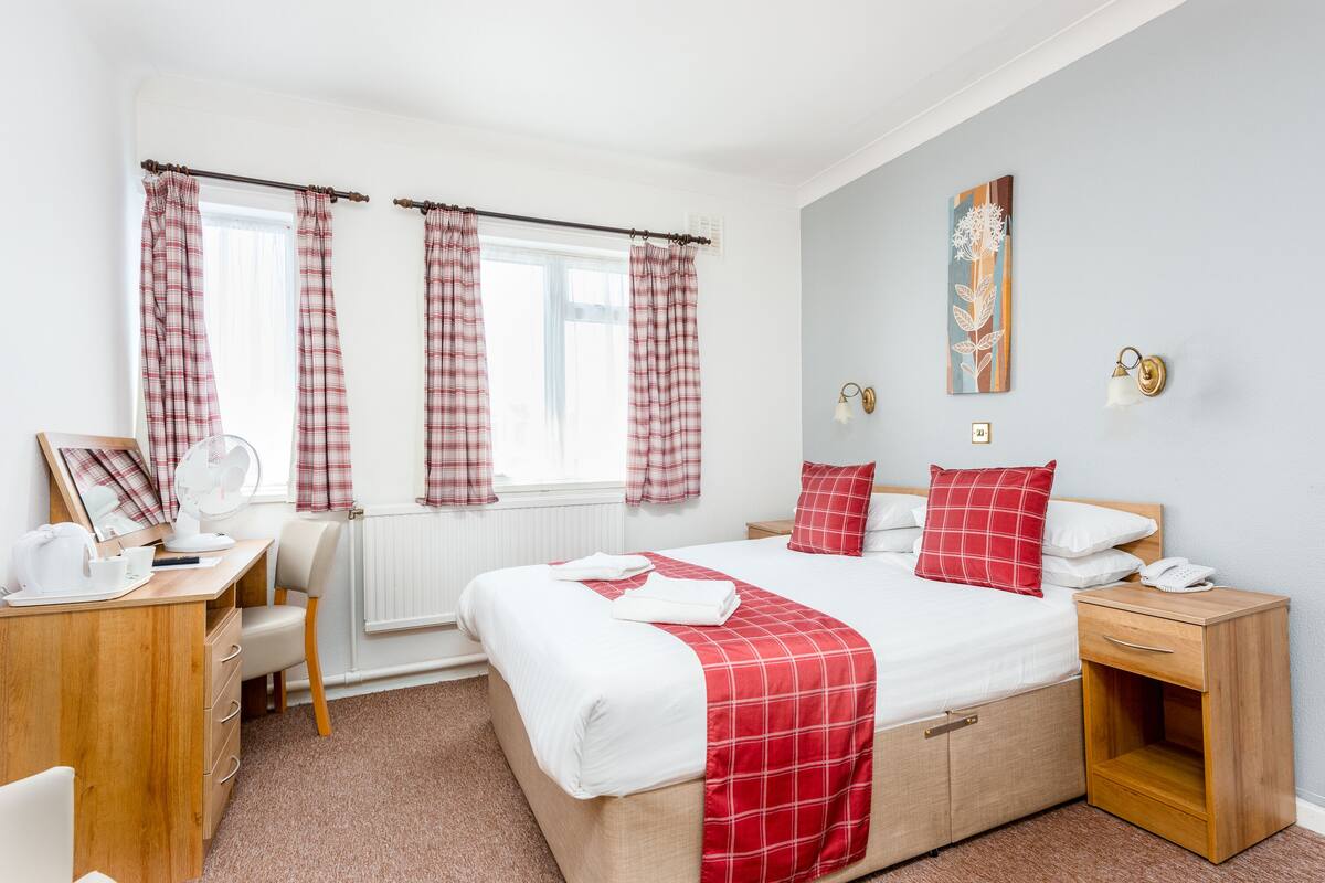 A bedroom with a bed, bedside table, desk, chair, and a window with curtains.