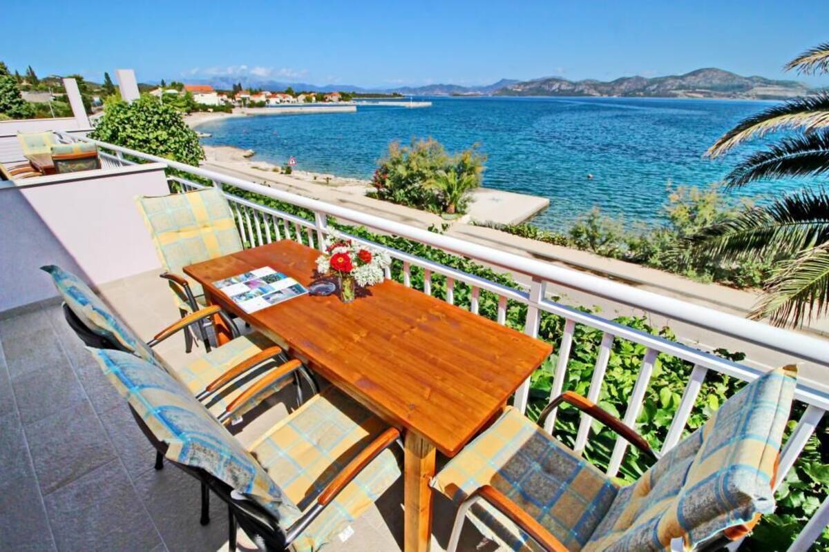 A wooden table with chairs and a bench on a balcony overlooking a coastal area with a clear view of the sea and distant mountains.
