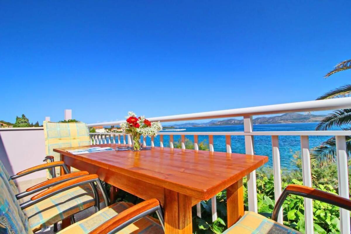 A wooden table with chairs and a vase of flowers on a balcony overlooking a body of water.