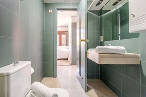 Een smalle badkamer met groene tegels, een wit toilet, een wastafel met een handdoek en een spiegel.