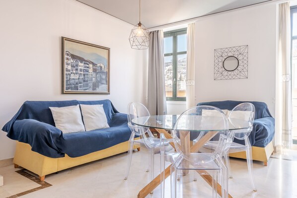 Un salon moderne avec un canapé bleu, une table à manger en verre et des chaises transparentes. Un tableau encadré est accroché au mur, et une œuvre d’art aux motifs géométriques est placée au-dessus de la fenêtre.
