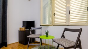 A room with a wooden floor, a television on a dark cabinet, two patterned chairs, a small green table with a plant, and a window with blinds. - The Point Room (Dubrovnik)