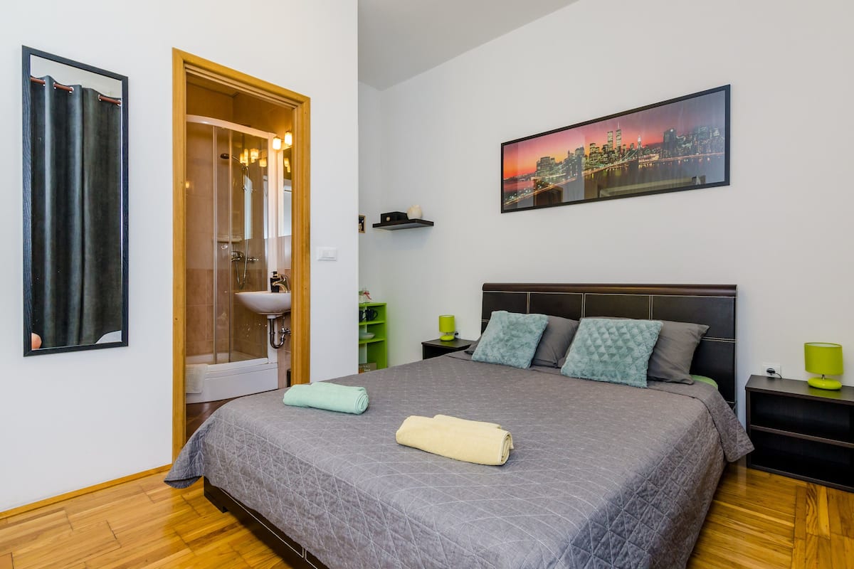 Un dormitorio con cama, mesita de noche, estante verde, baño con ducha y una pintura de paisaje urbano en la pared.