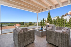 Un balcón con mobiliario de mimbre, una mesa de vidrio y vistas al mar y la vegetación.