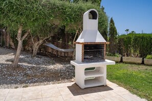 Una barbacoa exterior amb xemeneia, en un jardí amb arbres i una cadira espanyola.