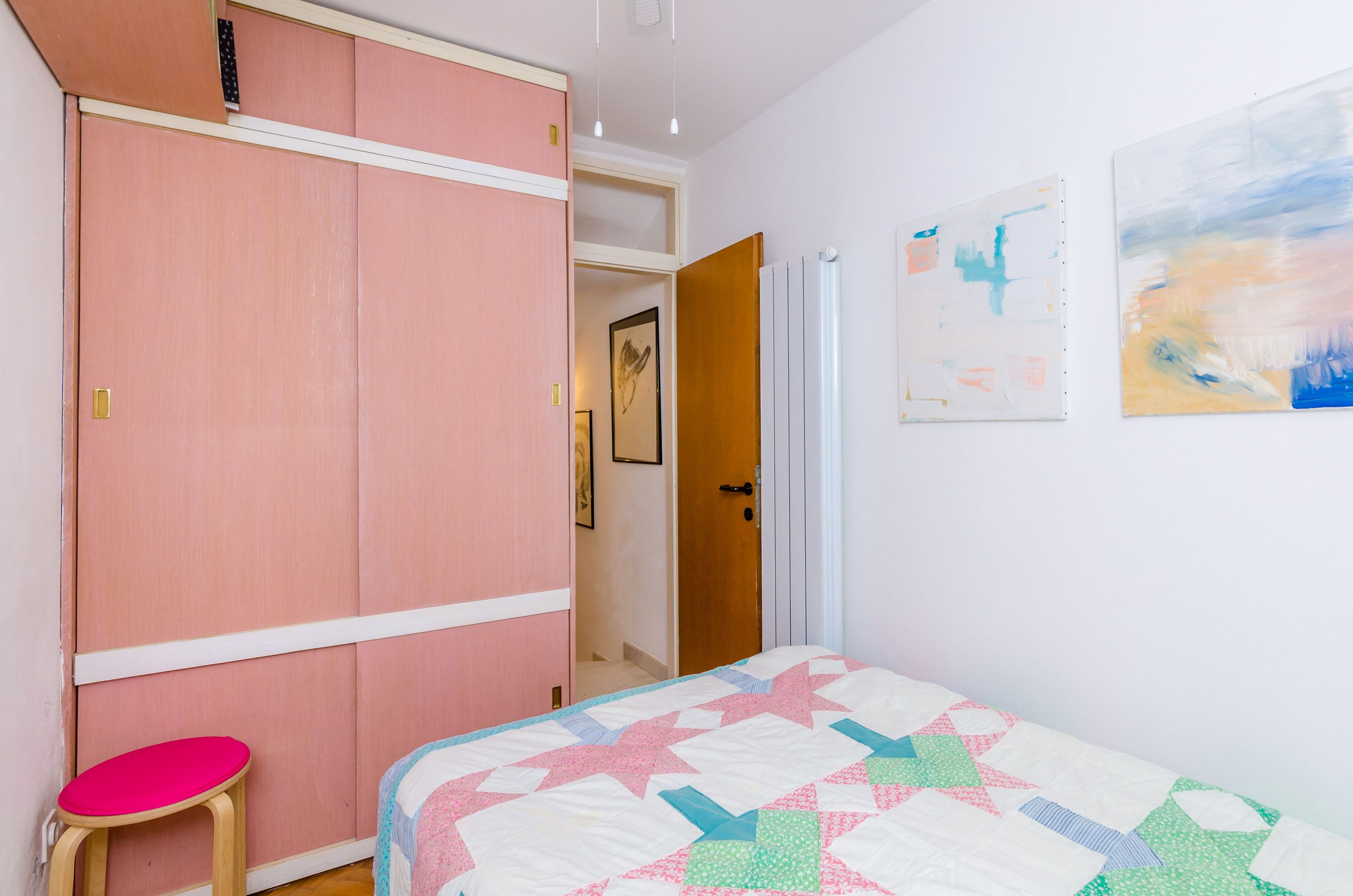 A bedroom with a pink wardrobe, a bed with a colorful quilt, and two abstract paintings on the wall.