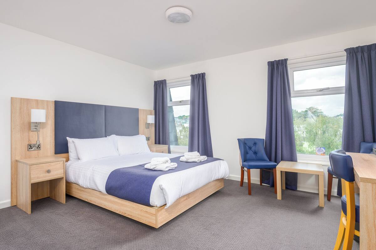 Ein Hotelzimmer mit Bett, Schreibtisch, zwei Stühlen, einem kleinen Tisch und einem fensterbepflanzten Fenster.