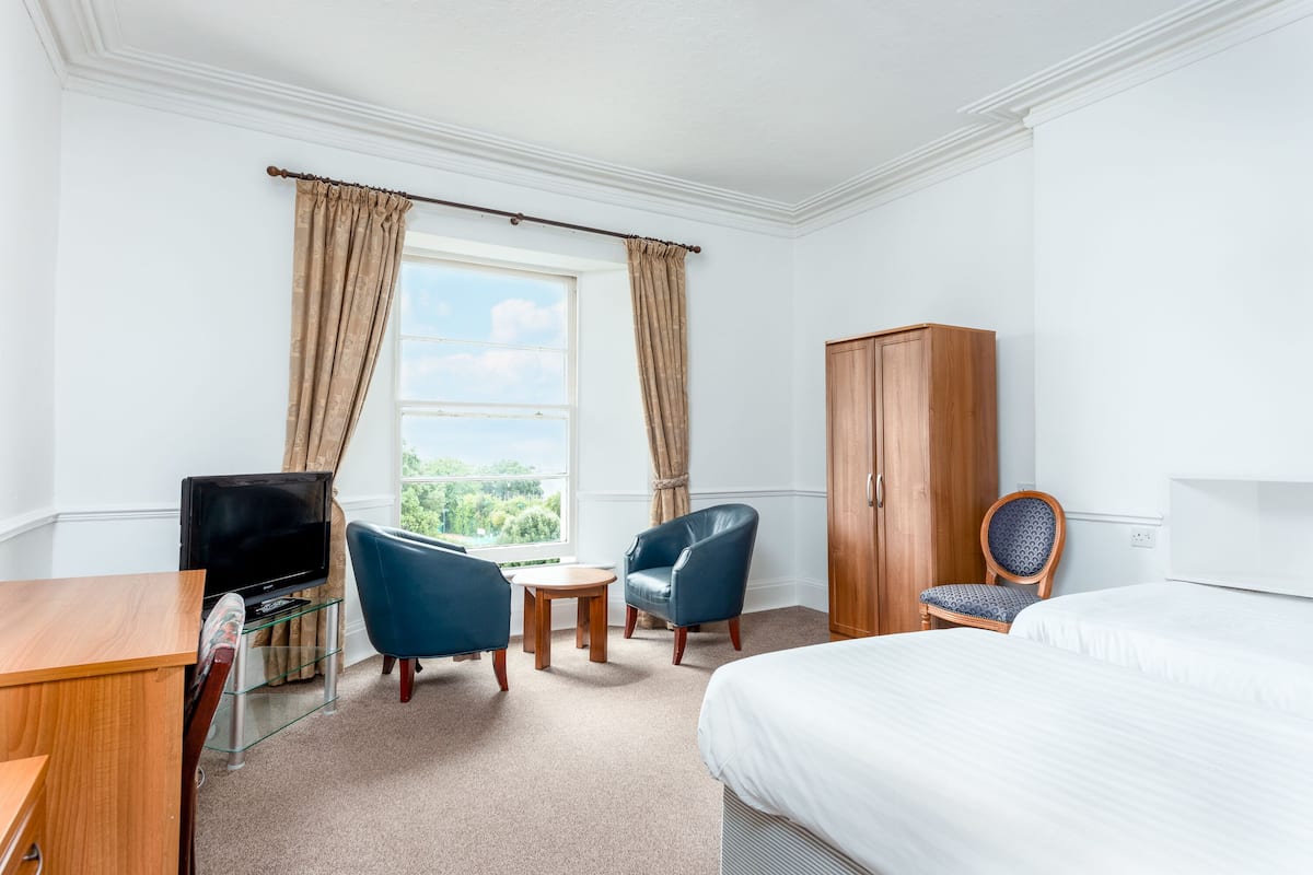 Ein Hotelzimmer mit Bett, Schreibtisch, Fernseher, zwei Stühlen, einem Kleiderschrank und einem Fenster mit Vorhängen.