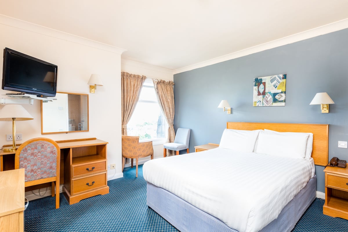 Ein Hotelzimmer mit Bett, Schreibtisch, Stuhl, Fernseher und einem Kunstwerk an der Wand.