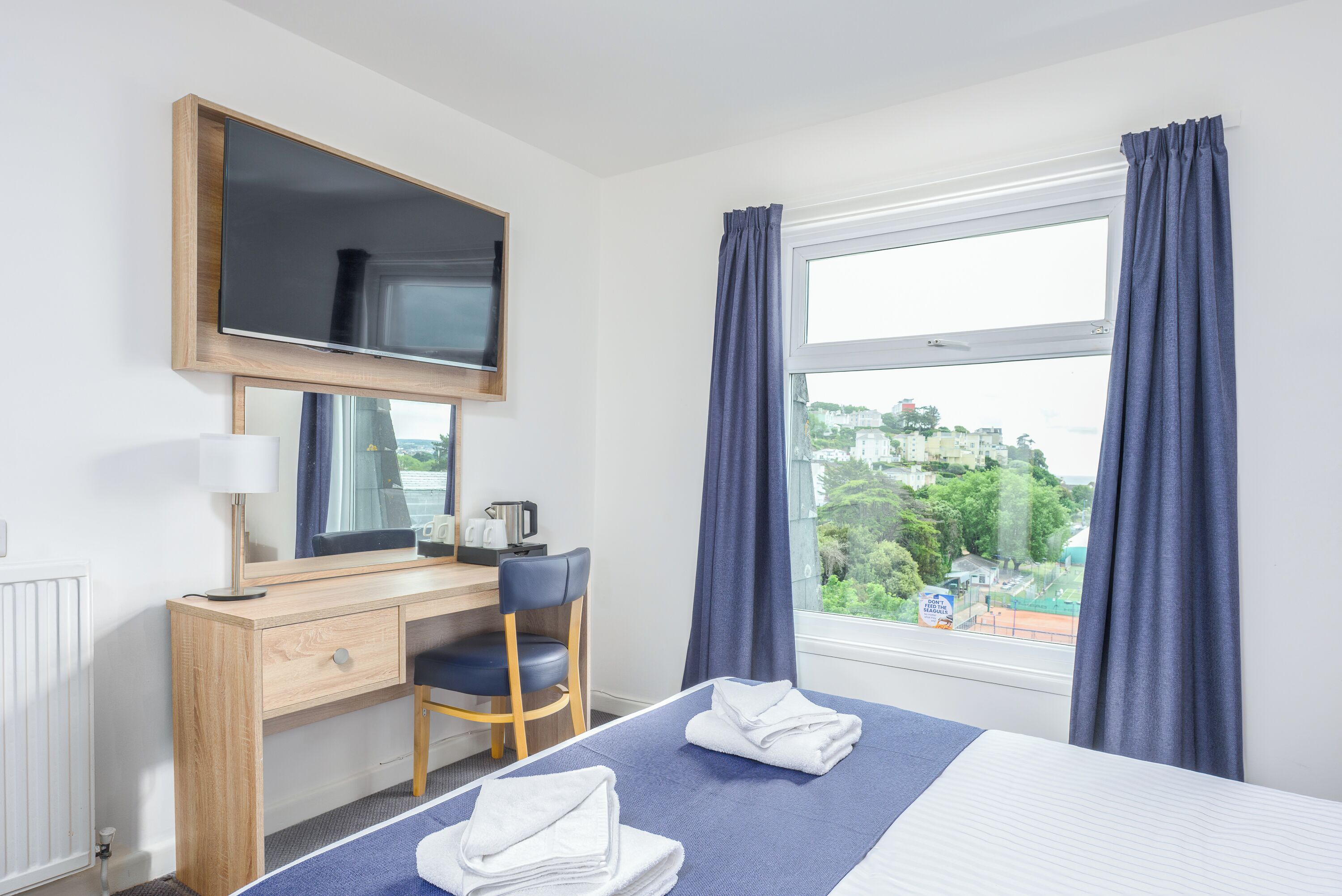 Une chambre d’hôtel comprenant un lit, un bureau avec une chaise, un miroir, une fenêtre avec des rideaux et une vue sur l’extérieur.
