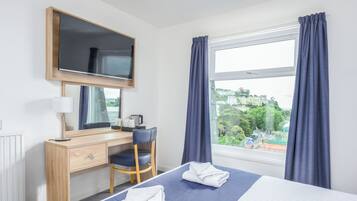 Une chambre d’hôtel comprenant un lit, un bureau avec une chaise, un miroir, une fenêtre avec des rideaux et une vue sur l’extérieur.