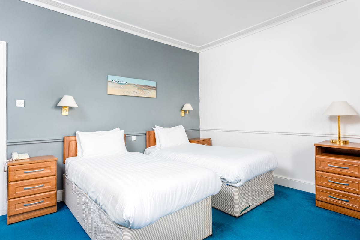 Ein Hotelzimmer mit zwei Betten, Holz-Nachttischen, einem blauen Teppich und Wandleuchten.