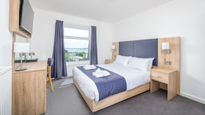 A hotel room with a bed, bedside table, TV, desk, and window with curtains.