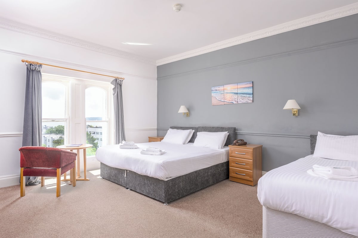Ein Hotelzimmer mit einem großen Bett, einem Nachttisch aus Holz, einem roten Sessel und Blick auf die Stadt durch das Fenster.