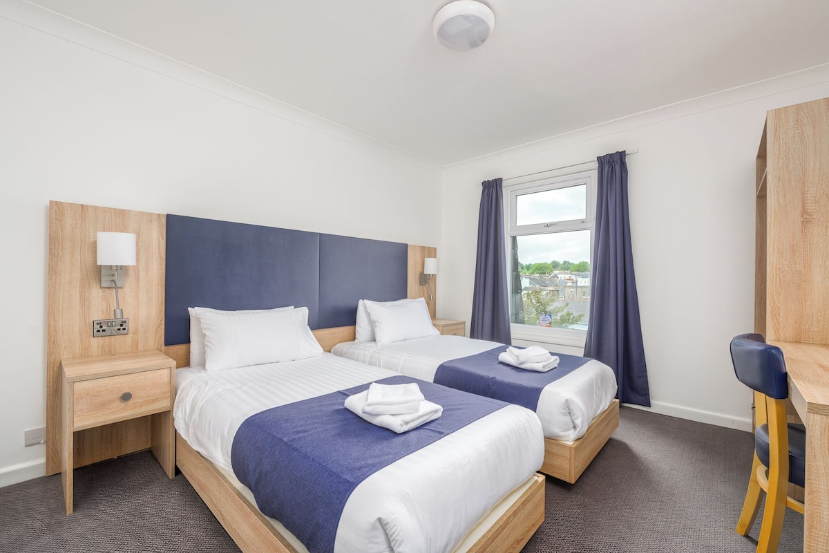 Ein Hotelzimmer mit zwei Betten, einem Schreibtisch und einem Fenster mit Vorhängen.