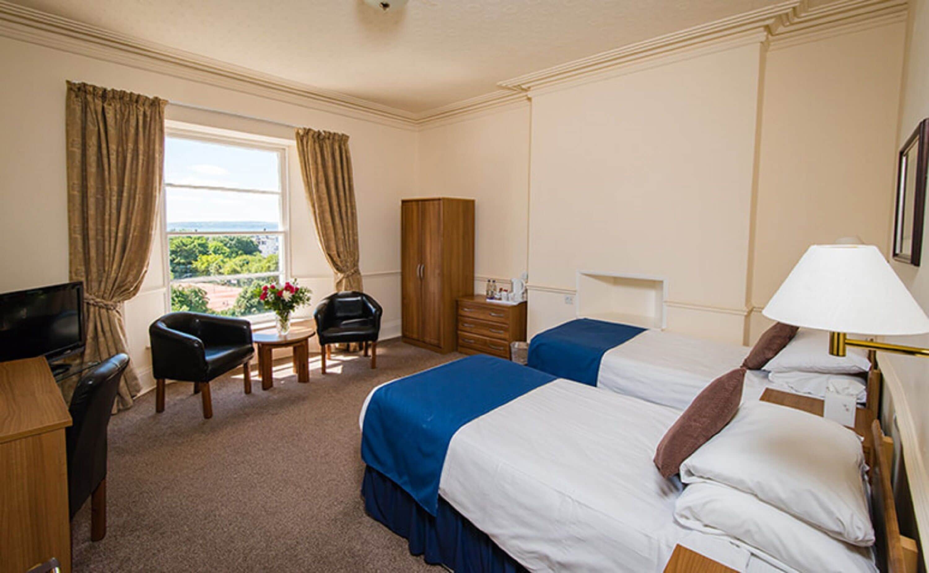 A hotel room with two beds, a desk, a TV, a wardrobe, and a window with curtains.