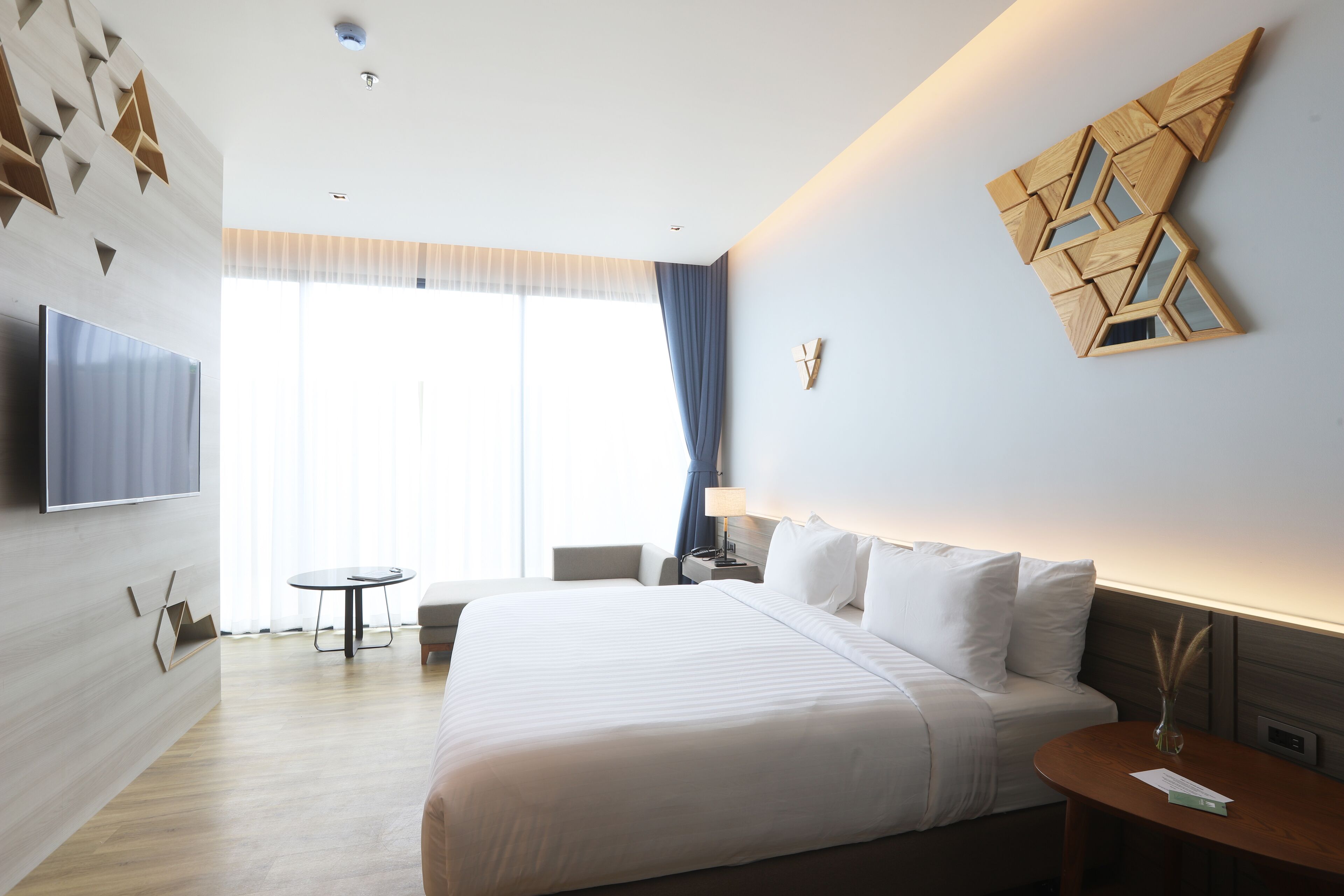A modern hotel room with a large bed, a flat-screen TV mounted on the wall, a small round table, and a window with curtains.