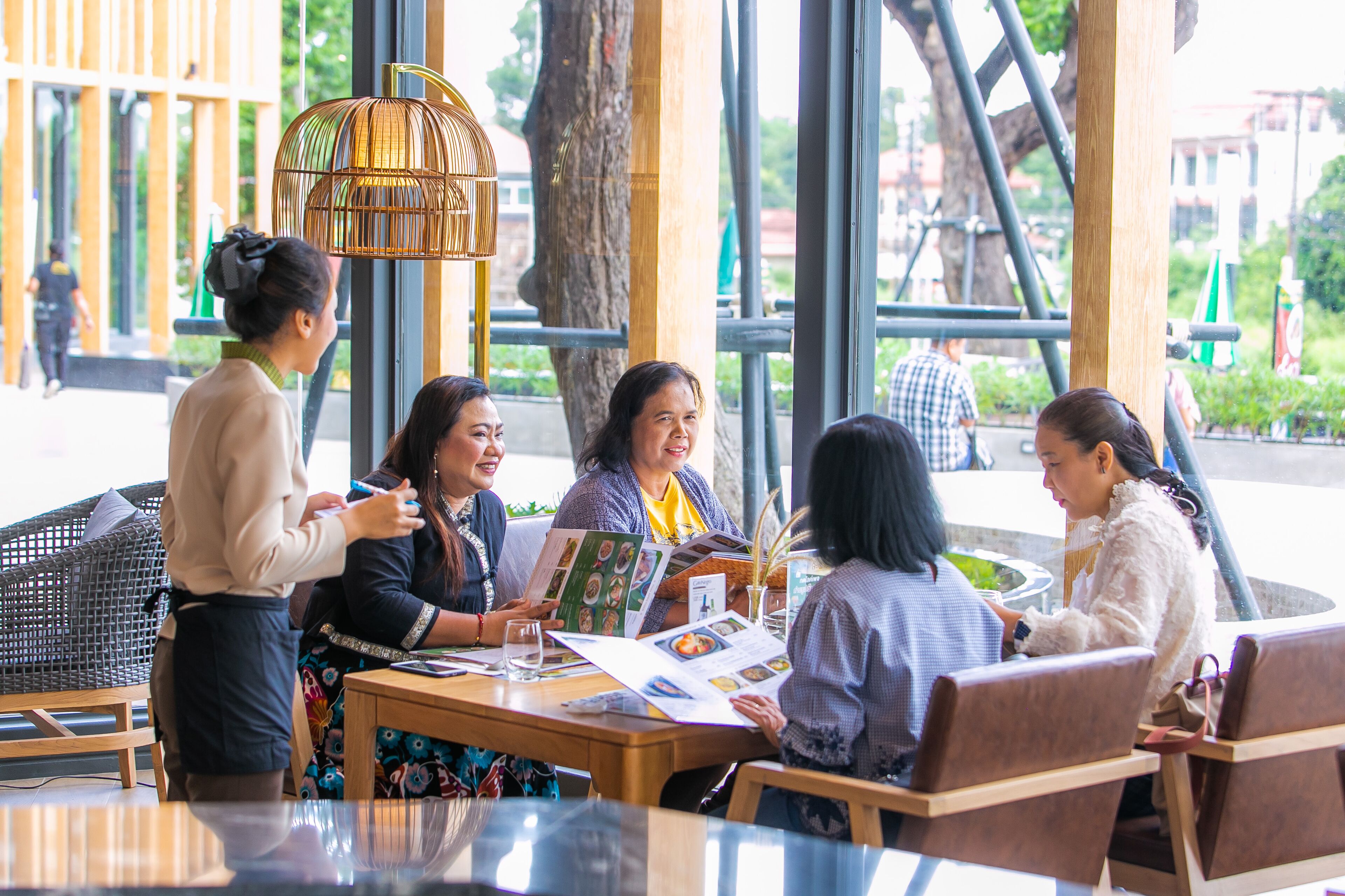 A group of people seated at a table with menus, engaged in conversation in a cafe with large windows and a tree outside.