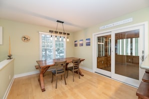 A dining area with a wooden table and chairs, a clock, and a sliding glass door leading to a wooden deck. - Tranquility Beach House - Beautiful cape cod style home just a short walk to Oak Street beach access (South Haven)