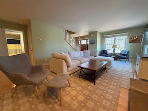 A living room with a sectional sofa, two armchairs, a coffee table, a staircase, and a window with a view. - Tranquility Beach House - Beautiful cape cod style home just a short walk to Oak Street beach access (South Haven)