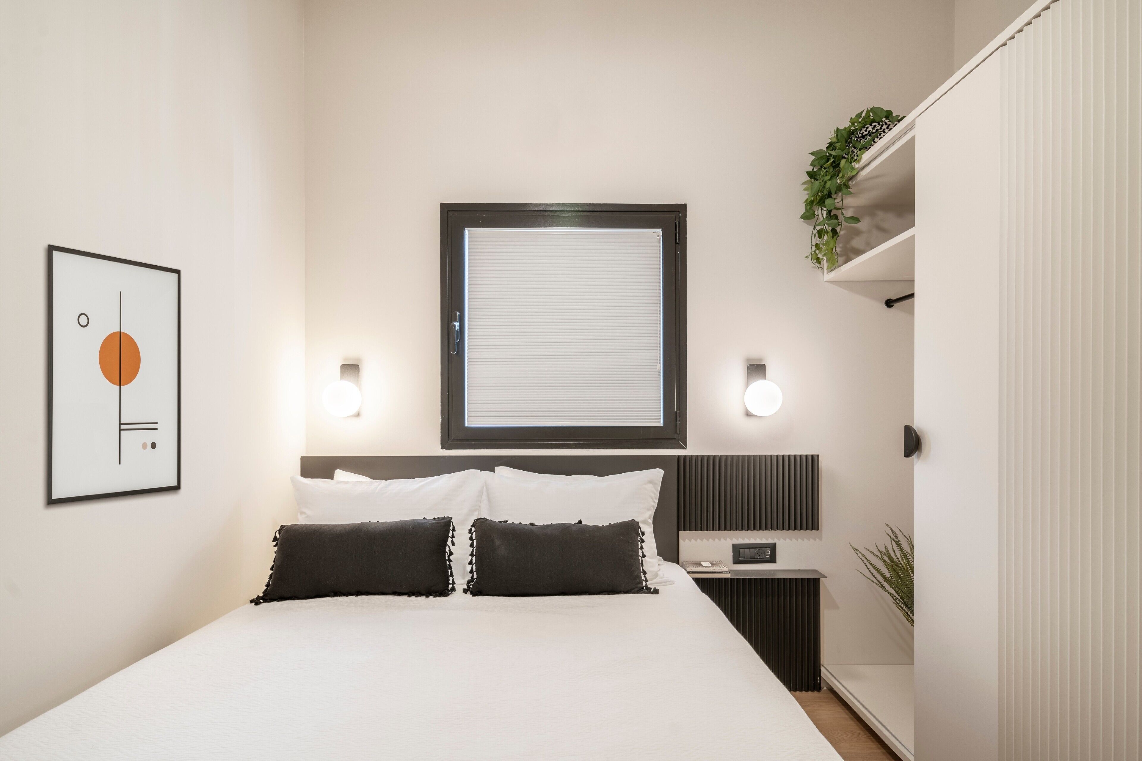 A modern hotel room with a large bed, a bedside table with a lamp, a framed artwork on the wall, and a window with blinds.