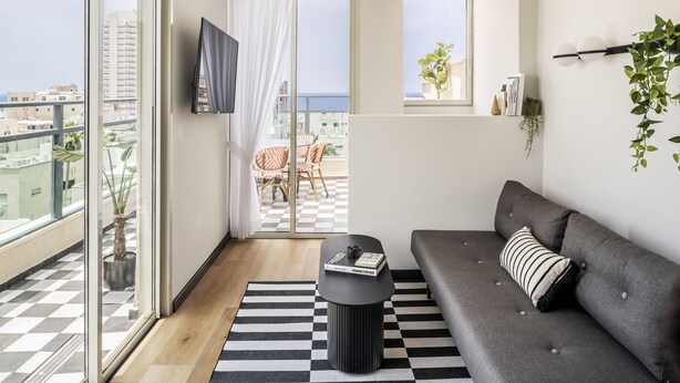 A modern living room with a grey sofa, a black coffee table, and a checkered rug. There is a balcony with a view of the city and a plant on the windowsill. - Mr. Montana - By TLV2GO (Tel Aviv)