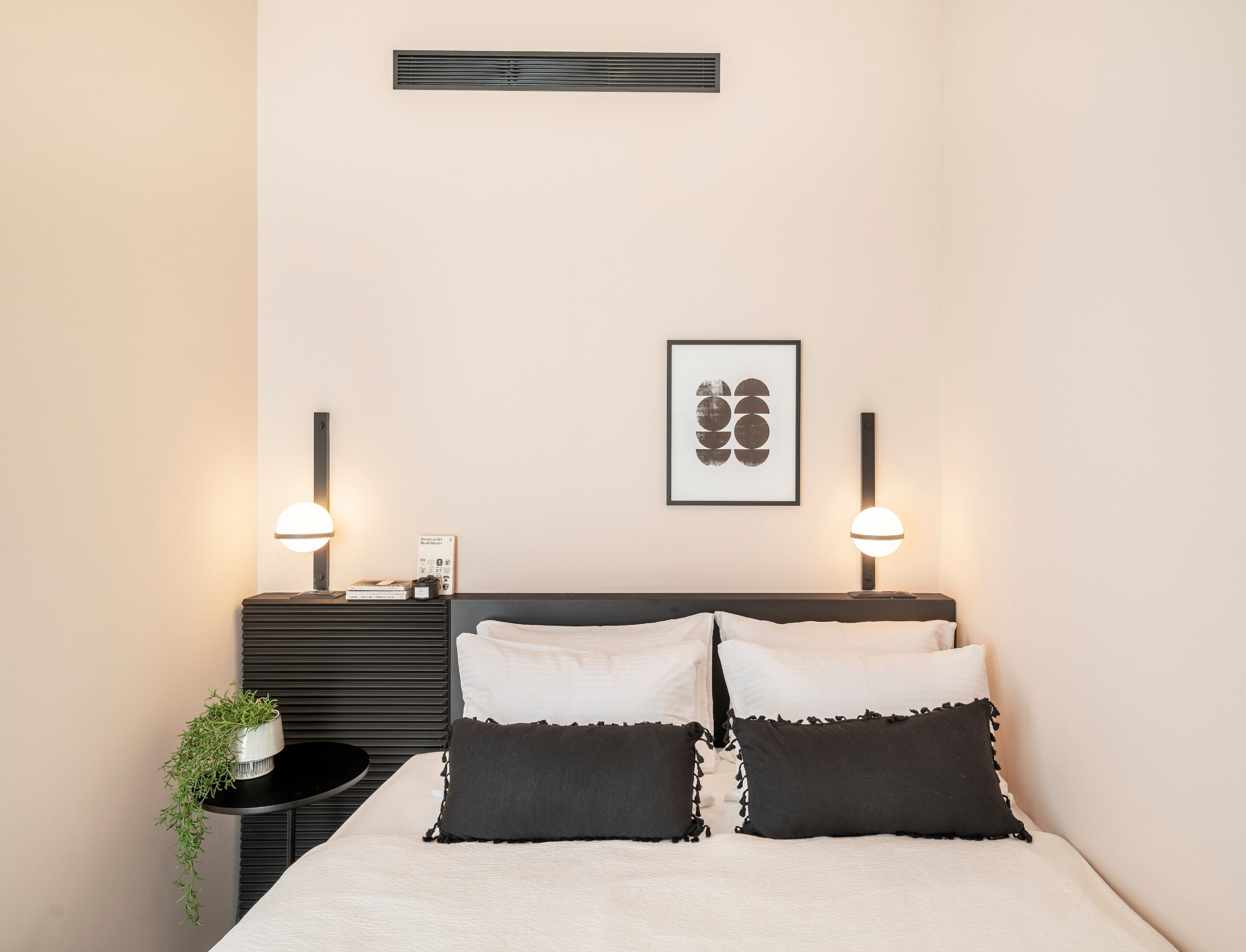Ein ordentlich eingerichtetes Schlafzimmer mit einem Bett, Nachttischlampen, einem gerahmten Kunstwerk und einem Tisch mit einer Pflanze.