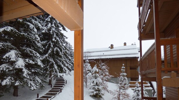 Ein Holzbalkon mit einem herzförmigen Ausschnitt, eine mit Schnee bedeckte Treppe, die zu einem Gebäude führt, und mit Schnee beladene Bäume.