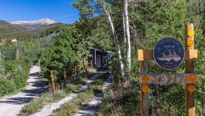 Uma cabana rústica com um caminho de terra que leva até ela, cercada por árvores e montanhas.