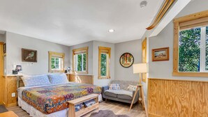 Quarto com uma cama grande, um sofá, cabeceira de madeira, janela com vista para árvores e um quadro na parede.